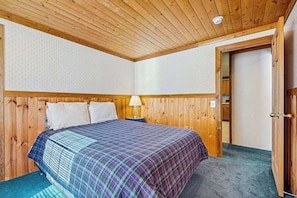 A bedroom with a wooden ceiling, a bed with a plaid bedspread, a nightstand with a lamp, and a door leading to another room. - Spacious 3BR Mountainview - Fireplace - Deck (Leavenworth)