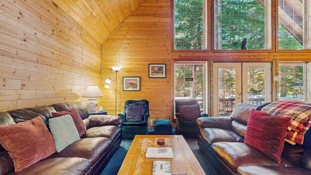 Ein gemütliches Blockhauss Innenleben mit Holzwänden, Ledersofas, einem Couchtisch und großen Fenstern mit Blick ins Freie.
