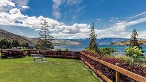 Uma área gramada com um banco, uma cerca de madeira e vista para um lago com montanhas ao fundo.