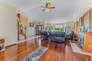A spacious living room with hardwood flooring, a ceiling fan, a fireplace, and a large window. - Spanish-style 6BR Mountainview - Deck - W&D (East Wenatchee)