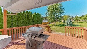 A covered outdoor deck with a barbecue grill, wooden railing, and a view of a green landscape. - Spanish-style 6BR Mountainview - Deck - W&D (East Wenatchee)
