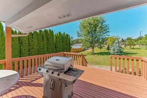 A covered outdoor deck with a barbecue grill, wooden railing, and a view of a green landscape. - Spanish-style 6BR Mountainview - Deck - W&D (East Wenatchee)