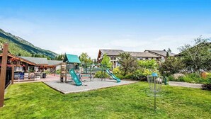 A playground with slides and a climbing structure, surrounded by a grassy area and buildings. - Downtown Studio 1st-Floor - Icicle Village Resort (Leavenworth)