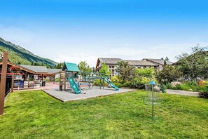 Ein Spielplatz mit Rutschen und einer Kletteranlage, umgeben von einer Rasenfläche und Gebäuden.