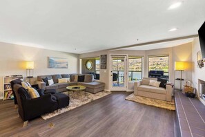 Ein geräumiges Wohnzimmer mit einem Ecksofa, einem schwarzen Sessel und einem Couchtisch. Zwei Fenster bieten einen Blick ins Freie.