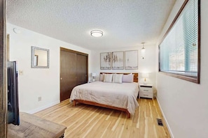Un dormitorio con piso de madera, una cama con cabecera, una mesita de noche, una lámpara, un espejo y una obra de arte en la pared.