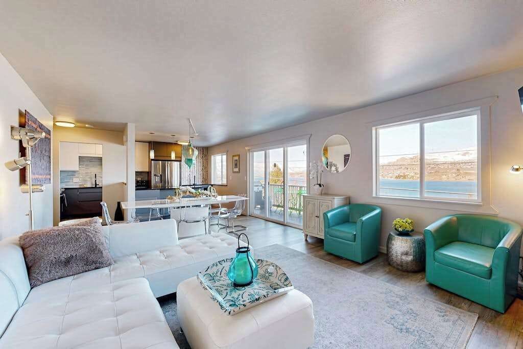 A modern living room with a white sofa, teal armchairs, and a dining area with a kitchen in the background.