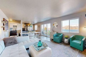 A modern living room with a white sofa, teal armchairs, and a dining area with a kitchen in the background.