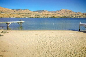 A sandy beach with a wooden dock extending into a calm lake, surrounded by hills.