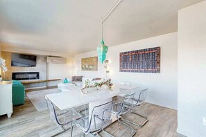 A modern living room with a white dining table, clear chairs, a teal pendant light, a flat-screen TV, and a bookshelf.