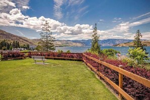 A grassy outdoor area with a wooden fence, a bench, and a view of a lake with mountains in the background. - Updated 3BR Lakeview - Balcony - Washer/Dryer (Chelan)