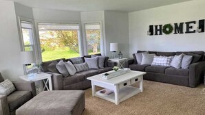 Ein Wohnzimmer mit zwei grauen Sofas, einem weißen Couchtisch und einem grauen Hocker. An der Wand hängt ein Schild mit der Aufschrift 'HOME' und ein Fenster bietet Blick auf Grünflächen.