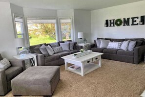 A living room with two grey sofas, a white coffee table, and a grey ottoman. There is a 'HOME' sign on the wall and a window with a view of greenery. - 4BR family friendly house with pool, hot tub, W&D (East Wenatchee)