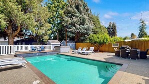 Ein rechteckiger Pool mit einer gefliesten Kante, umgeben von einer Holzterrasse. Liegestühle und ein Grillbereich befinden sich dahinter, mit einem Holzzaun im Hintergrund.
