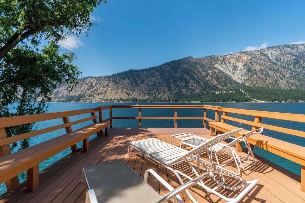A wooden deck with benches and lounge chairs overlooking a lake and mountains.