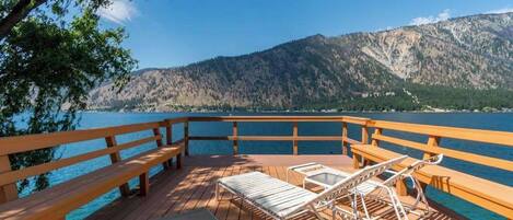A wooden deck with benches and lounge chairs overlooking a lake and mountains.