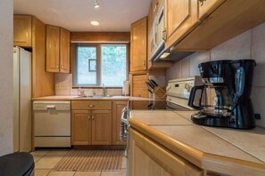 A compact kitchen with wooden cabinets, a dishwasher, a coffee maker, and a window with blinds.