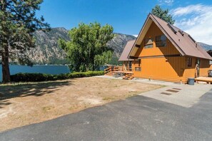 Une cabane en bois, dotée d’un toit en pente, située au bord d’un plan d’eau, avec des montagnes en arrière-plan.