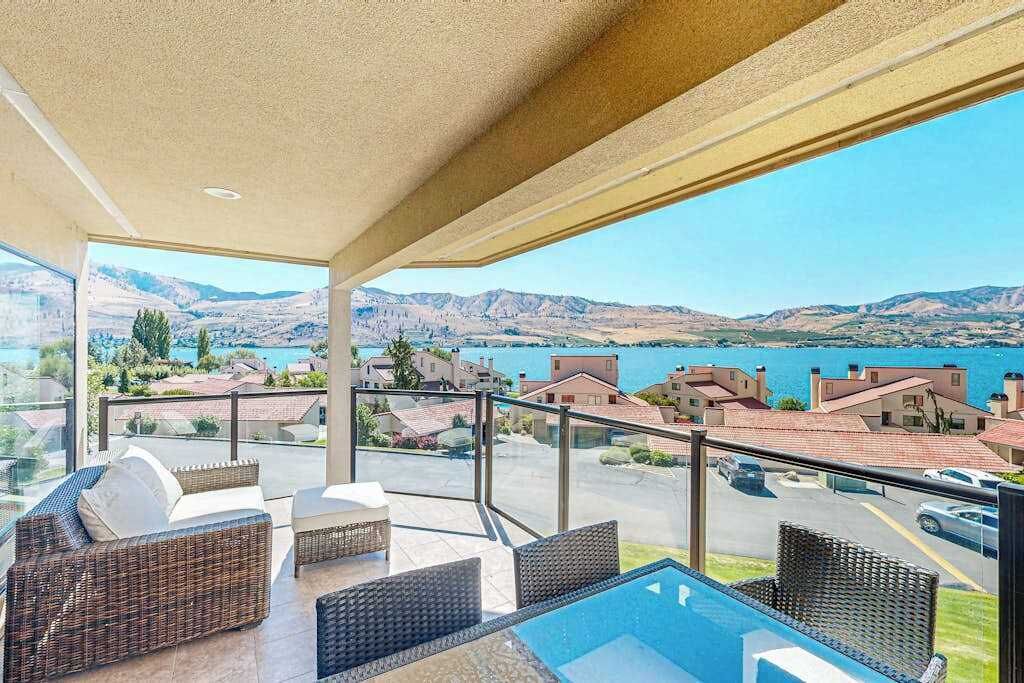 A balcony with wicker furniture, a glass table, and a view of a lake and mountains.