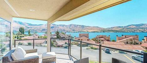 A balcony with wicker furniture, a glass table, and a view of a lake and mountains.