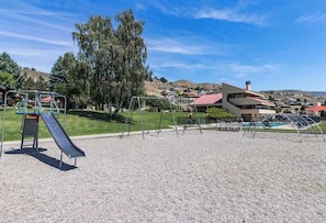 A playground with a slide and swings, surrounded by a gravel area, with houses and hills in the background. - 2BR lakeside condo with shared pools and hot tub (Chelan)