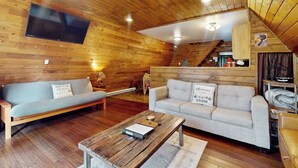 Ein gemütliches Blockhaus-Interieur mit Holzbalkendecke, einem wandmontierten Flachbildfernseher, einem Sofa, einem Couchtisch und einer Couch.