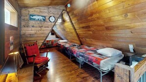 Eine Hütte mit geneigtem Dach, Holzwänden, einem Bett mit gemustertem Überwurf, einem roten Sessel und einem kleinen Tisch mit einer Lampe.