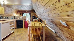 Ein gemütliches Blockhaus-Interieur mit einem Holzespeisetisch, Stühlen, einer Küchenzeile mit Spüle und Geräten sowie einem Bücherregal.