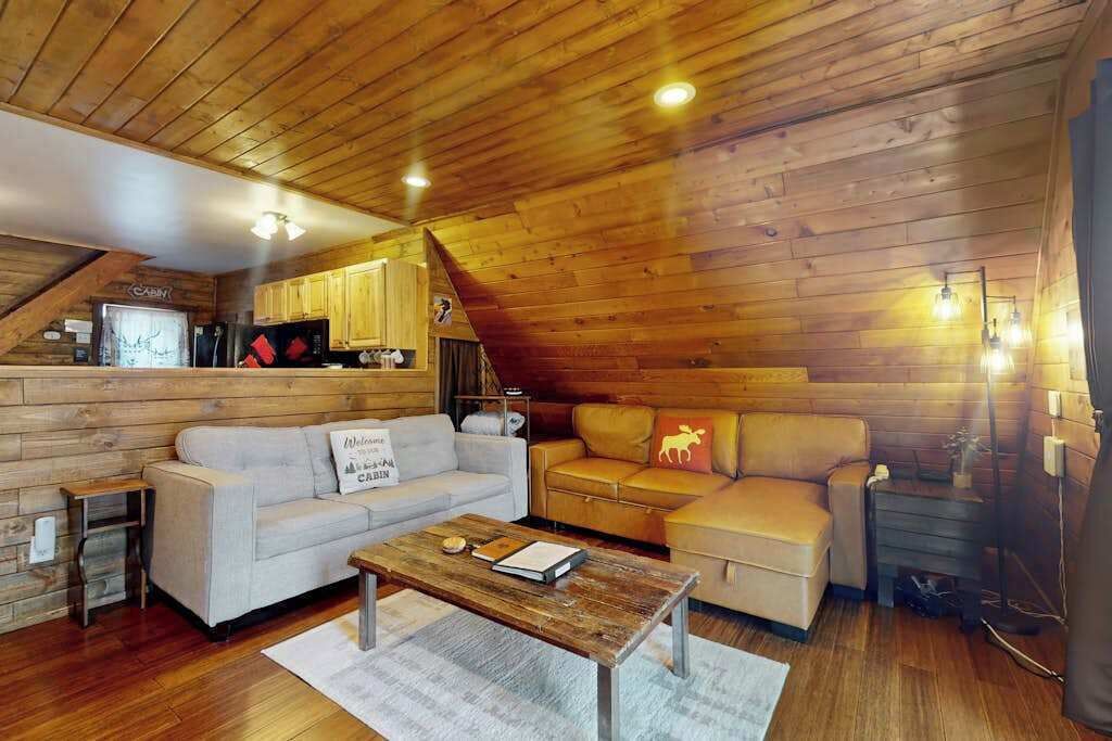 A cozy cabin interior with a wooden ceiling, a sofa, a coffee table, and a kitchenette.
