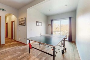 A room with a ping pong table, wooden flooring, a balcony with a view, and a door leading to another room.