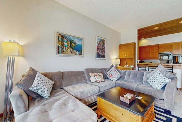 A modern living room with a sectional sofa, a coffee table, and a kitchen area in the background. - Charming 2BR Lakefront - Patio - Pool (Chelan)
