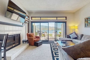A living room with a fireplace, a sofa, a chair, and a view of a balcony with outdoor furniture. - Charming 2BR Lakefront - Patio - Pool (Chelan)