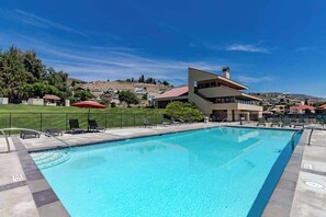 A swimming pool with a modern building in the background, a grassy area with lounge chairs, and a clear blue sky. - Charming 2BR Lakefront - Patio - Pool (Chelan)
