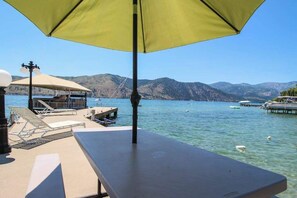 Ein Steg am See mit Tisch und Stühlen, ein überdachter Bereich mit einer Lampe und Blick auf Berge und Boote.