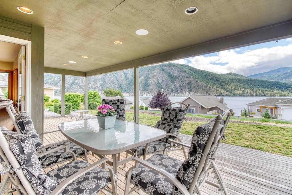 A covered outdoor seating area with a table and chairs, overlooking a lake and mountains.