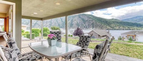 A covered outdoor seating area with a table and chairs, overlooking a lake and mountains.