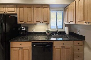 Eine Küche mit Holzschränken, einem schwarzen Kühlschrank, einer Spülmaschine, einem Spülbecken und einem Fenster mit Jalousien.