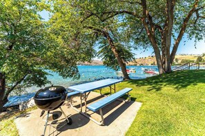 A picnic table with a grill, a bench, and a view of a lake with boats.