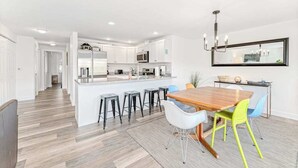 A modern kitchen with white cabinets, a central island with stools, and a dining area with a wooden table and colorful chairs. - Bright 2BR Lakeview - Deck - Pool - (Chelan)