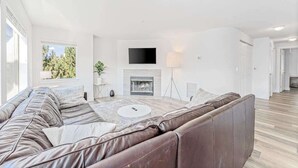 A modern living room with a leather sofa, a fireplace, a flat-screen TV, and a large window. - Bright 2BR Lakeview - Deck - Pool - (Chelan)