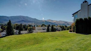 A residential area with a grassy lawn, a modern house, and mountains in the background. - Downtown 2BR - Patio - Pool - Washer/Dryer (Chelan)