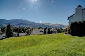 A residential area with a grassy lawn, a modern house, and mountains in the background. - Downtown 2BR - Patio - Pool - Washer/Dryer (Chelan)