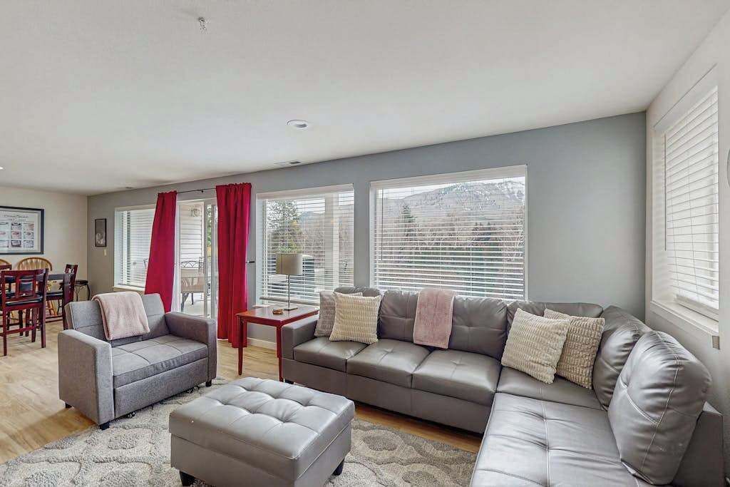 A living room with a grey sofa, a grey armchair, and a matching ottoman. There are two windows with blinds, a dining table with chairs, and a red curtain.