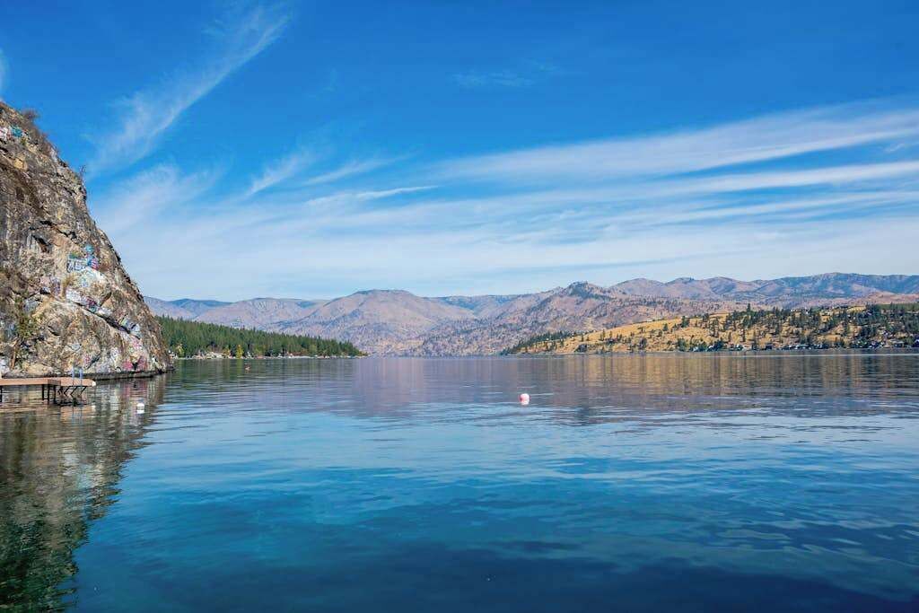 Un llac tranquil amb riba rocosa i muntanyes llunyanes sota un cel serè.