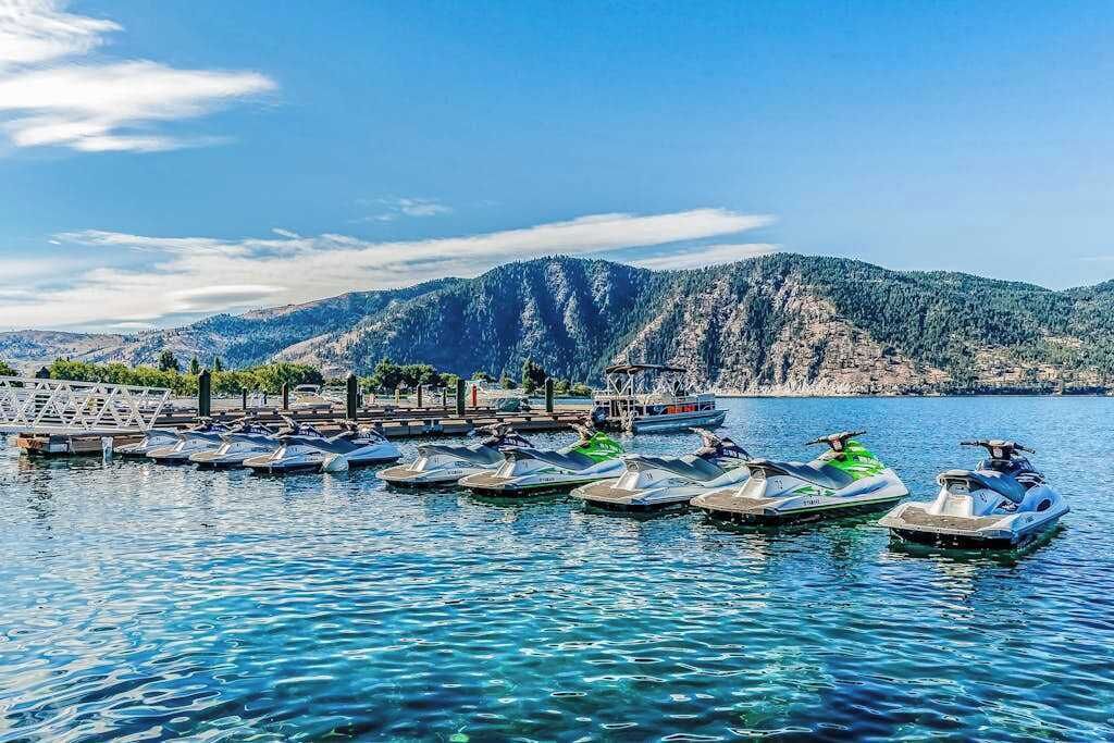 En lystbådehavn med flere både og jetski ved kaj, omgivet af bjerge og krystalklart vand.