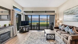 A living room with a fireplace, a television, a sofa, and a view of the outdoors. - Comfy 1BR Lakefront - Patio - Pool - (Chelan)