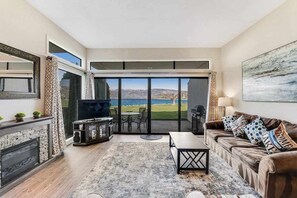 A living room with a fireplace, a television, a sofa, and a view of the outdoors. - Comfy 1BR Lakefront - Patio - Pool - (Chelan)