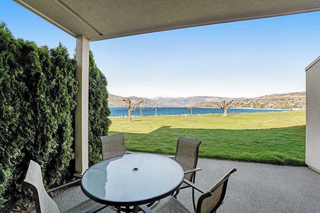 A covered patio with a round table and chairs, overlooking a grassy area and a body of water with trees and hills in the distance.