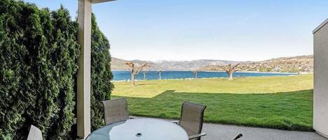A covered patio with a round table and chairs, overlooking a grassy area and a body of water with trees and hills in the distance.