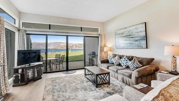 A living room with a television, sofa, coffee table, and a view of a lake through large sliding glass doors. - Comfy 1BR Lakefront - Patio - Pool - (Chelan)
